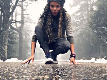 Portrait of beautiful woman crouching on street amidst trees during winter