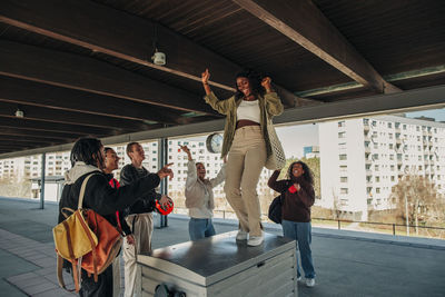 Multiracial friends cheering while woman dancing on seat at railroad station platform