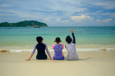 Rear view of female friends sitting on shore at beach against sky