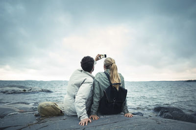 Couple taking selfie while sitting on rock by sea