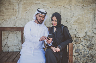Couple wearing traditional clothing using smart phone while sitting on bench
