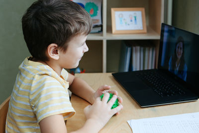 8 years old boy sit by desk with laptop and do exercise with massage ball. side view.