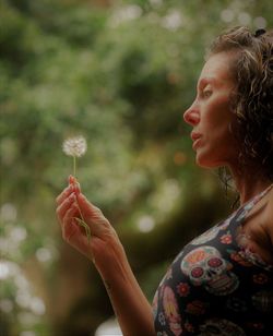 Side view of woman holding dandelion against trees