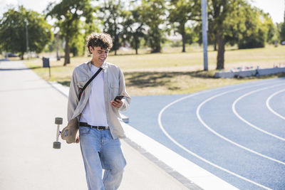 Smiling man holding skateboard using smart phone walking on footpath
