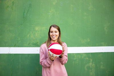 Portrait of smiling young woman standing with ball against wall