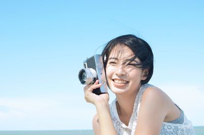 Smiling young woman holding camera against clear sky