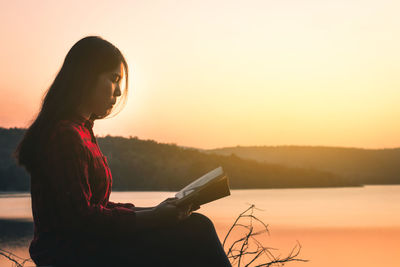 Woman sitting by lake against sky during sunset