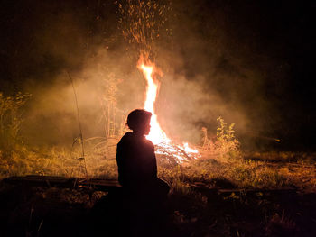 Silhouette boy sitting by campfire on field at night