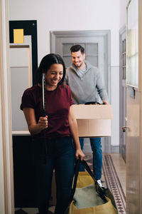 Smiling couple carrying box and bag while walking in corridor at home