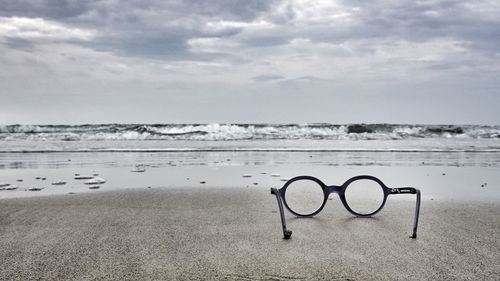Close-up of eyeglasses on shore at beach against cloudy sky