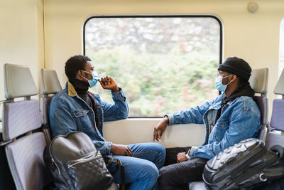 Side view of calm black men in medical masks sitting on passenger seats and looking out of window while traveling by train during covid 19 epidemic