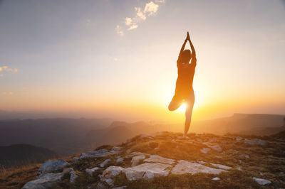 Silhouette of a woman in a tree pose, yoga asana, stands near a cliff in the mountains against the