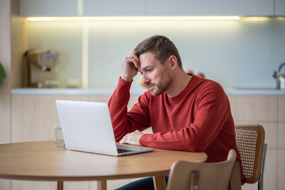 Jobless upset man sitting at table with laptop computer, looking through vacancies, hunting for job