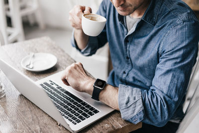 Mature man drinking coffee while working at laptop, looking at smartwatch