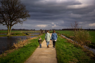 Rear view of women with girl walking on footpath against cloudy sky