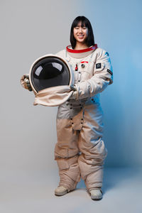 Young asian woman in spacesuit holding helmet and looking at camera under blue light before mission against gray background