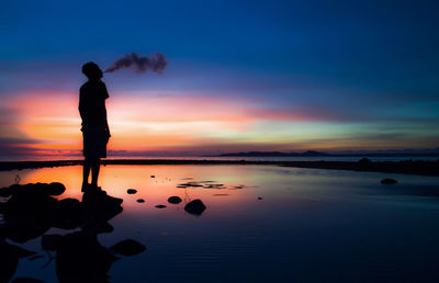 Silhouette man exhaling smoke at beach against sky during dusk