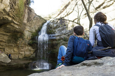 Back view of two young teenagers sitting on rock against waterfall