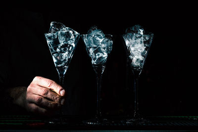 Close-up of human hand holding martini glasses with ice cubes against black background