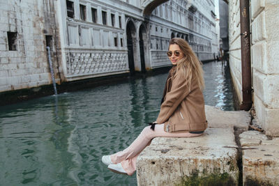 Side view of young woman sitting on retaining wall by canal in city