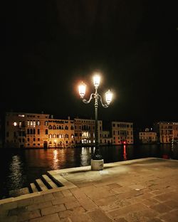 Illuminated street by buildings against sky at night