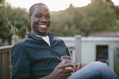 Portrait of smiling mid adult man holding glass while sitting at porch