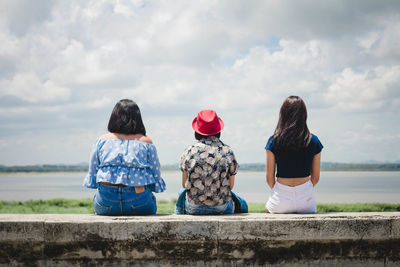 Rear view of women sitting on retaining wall against sky