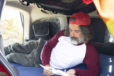 Bearded man in cap sitting in car trunk and reading book during trip picnic person