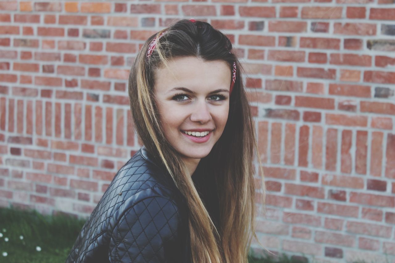 young adult, portrait, looking at camera, person, young women, front view, lifestyles, long hair, headshot, smiling, casual clothing, leisure activity, brick wall, wall - building feature, brown hair, black hair, standing