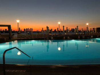 Illuminated street light reflecting on swimming pool against sky during sunset