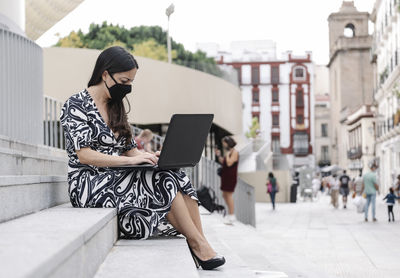 Full length of businesswoman wearing mask using laptop while sitting on steps outdoors