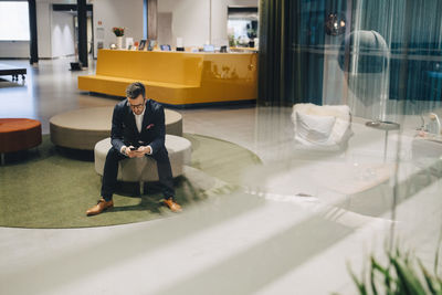 Full length of businessman using mobile phone while sitting in lobby at office