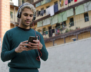 Young man with headphones holding smartphone in hand and listening to music in city