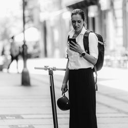 Businesswoman standing and using her smart phone with electric scooter next to her in the city.