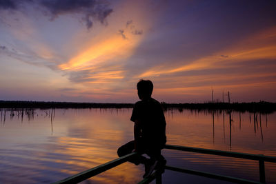 Rear view of silhouette man standing by lake against sky during sunset