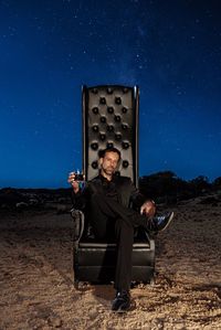 Portrait of mature man holding drink while sitting on chair against sky at night