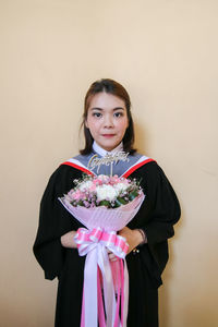 Portrait of young woman wearing graduation gown holding bouquet against wall