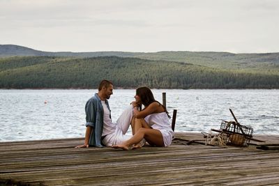 Romantic couple sitting on pier by lake against sky