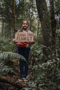 Ethnic kid raising cardboard piece with save the planet inscription while looking at camera in green forest