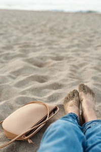 Close-up of bare tanned woman's feet covered with sand and a handbag on a sandy beach. 
