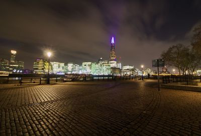 Illuminated city street by buildings against sky at night