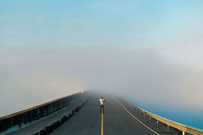 Rear view of man standing on bridge against sky during foggy weather