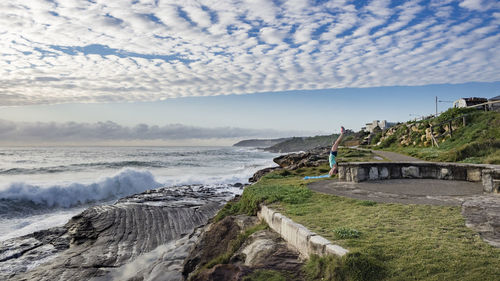 Person exercising on cliff by sea against cloudy sky