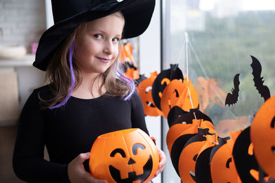 Little girl in costume of witch holding pumpkin jack with candies, celebrating halloween at home