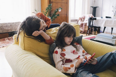 Girl using smart phone while sister leaning on sofa at home