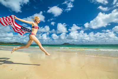 Side view of woman holding american flag while jumping at beach against cloudy sky