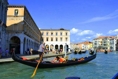 Man rowing gondola in canal against buildings and sky on sunny day