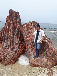 Full length of young man standing against brick wall at beach against sky