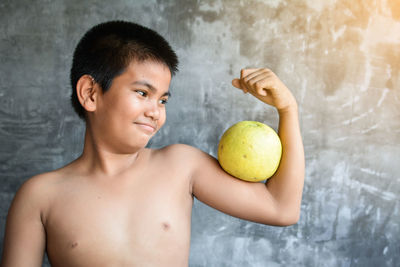 Shirtless boy holding food while sitting against wall