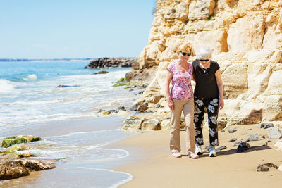 Two elderly women are walking along the rocky shore